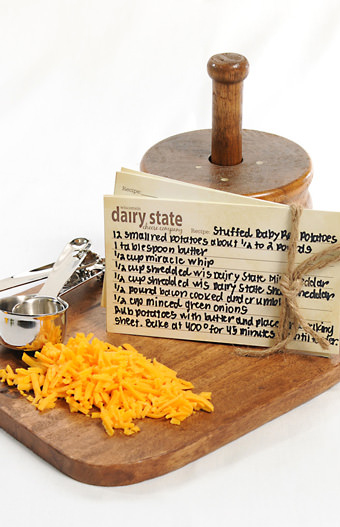 metal measuring cups, shredded cheddar and recipe cards on a wooden cutting board